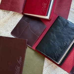 Puede incluir: Tres fundas rectangulares de cuero para cuadernos en color burdeos, negro y marrón oscuro. Cada funda presenta el texto en cursiva "With Love" grabado en la parte frontal. Las fundas están abiertas, mostrando bolsillos y ranuras interiores.