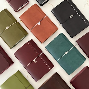 Könnte beinhalten: Eine Auswahl an Leder-Journals in verschiedenen Farben, darunter Burgunderrot, Olivgrün und Braun. Einige Journals haben dekorative Metallakzente und Verschlüsse, wie Nieten, Anhänger und Gummibänder. Die Journals sind auf einer hellen Oberfläche angeordnet.