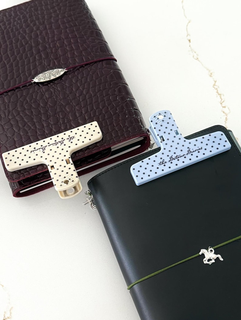 May include: A dark burgundy crocodile-textured notebook with a cream-colored polka dot clip and a black notebook with a light blue polka dot clip. Both clips feature the text "the detour diaries" printed in a cursive script on the front surface.