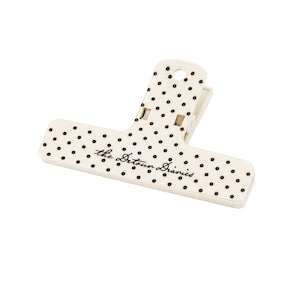 May include: A cream-colored plastic binder clip featuring a pattern of small black polka dots across its surface. The text "the Detour Diaries" is printed in a dark brown cursive script across the center of the wide, flat clip handle.