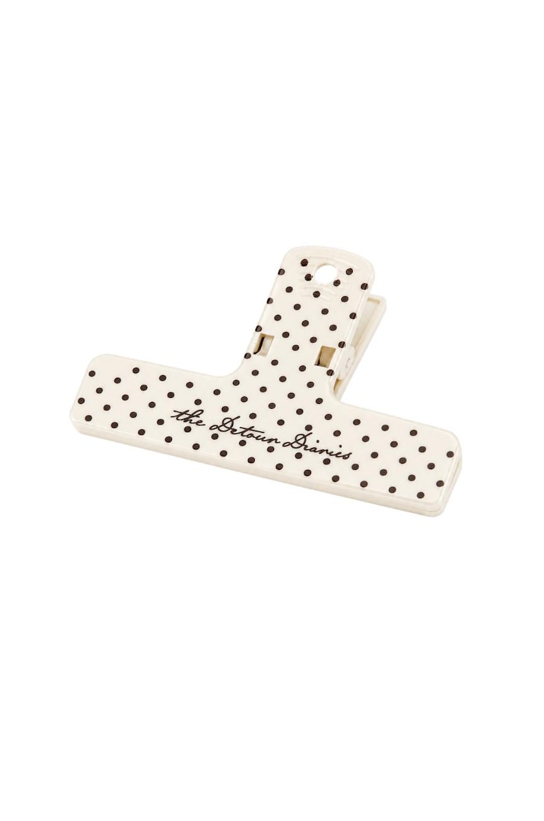 May include: A cream-colored plastic binder clip featuring a pattern of small black polka dots across its surface. The text "the Detour Diaries" is printed in a dark brown cursive script across the center of the wide, flat clip handle.