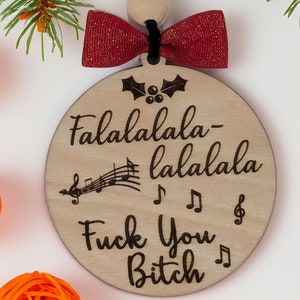 May include: Wooden Christmas ornament with the text "Falalalala-lalala Fuck You Bitch" and musical notes. The ornament has a red bow and black string for hanging. Decorative orange spheres and pine branches surround the ornament.