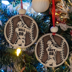 May include: Two baseball-themed Christmas ornaments hanging on a decorated tree. Each round ornament features a baseball player silhouette with a name and number, set against a baseball design. They hang from red ribbons.