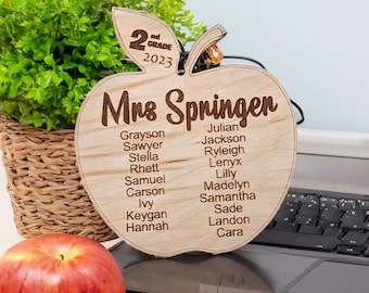 Może przedstawiać: Drewniana tabliczka w kształcie jabłka z tekstem "Mrs Springer" i "2nd Grade 2023" u góry. Tabliczka zawiera listę imion uczniów z klasy drugiej.