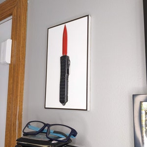 May include: A black and red knife with a black handle is displayed in a white frame with a black border. The knife is mounted on a wall.