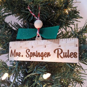 May include: A wooden ruler ornament with the text "Mrs. Springen Rules" and the year "2023" on it. The ornament is decorated with a green bow and a white bead.