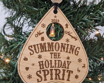 May include: Wooden Christmas ornament shaped like a Ouija board with the text "Summoning the Holiday Spirit" and a small light bulb in the center. The ornament is hanging on a Christmas tree.
