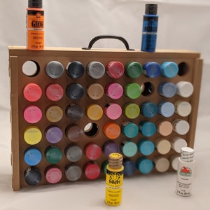 Puede incluir: Un organizador de pintura de madera con asa, que contiene una variedad de botellas de pintura de diferentes colores. El organizador tiene 48 ranuras para botellas de pintura. Las botellas de pintura son de 2 oz (59 ml).
