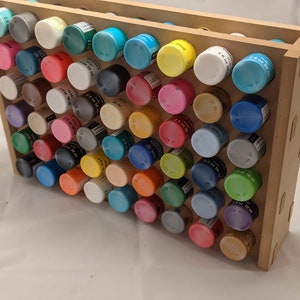 Puede incluir: Un estante de pintura de madera con 50 peque&ntilde;os frascos de pintura de varios colores. Los frascos est&aacute;n dispuestos en filas y columnas, y el estante est&aacute; hecho de madera marr&oacute;n claro.