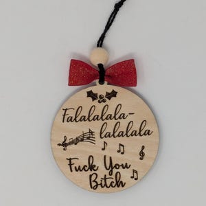 May include: A round wooden Christmas ornament with the text "Falalalala-lalala Fuck You Bitch" and musical notes. It has a red bow and a black string for hanging. The ornament is a humorous holiday decoration.