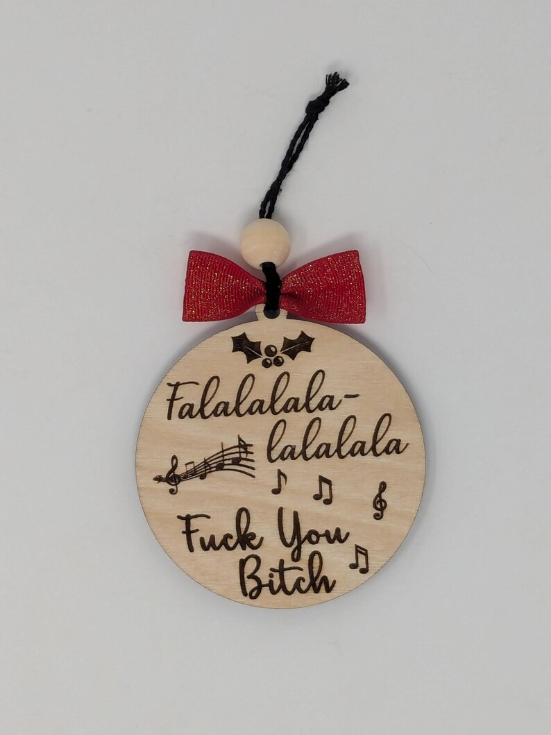 May include: A round wooden Christmas ornament with the text "Falalalala-lalala Fuck You Bitch" and musical notes. It has a red bow and a black string for hanging. The ornament is a humorous holiday decoration.