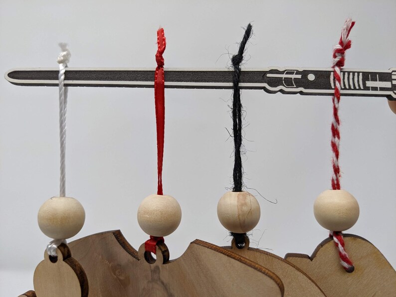 May include: Wooden ornaments with a lightsaber design, each suspended by a different colored string. The strings are white, red, black, and red-and-white striped. The ornaments are attached to a wooden base.