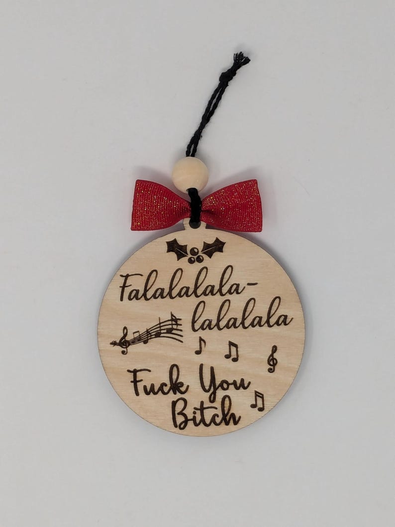 May include: A round wooden Christmas ornament with the text "Falalalala-lalala Fuck You Bitch" in a black font. The ornament has a red bow and a black string for hanging. The ornament is a humorous holiday decoration.
