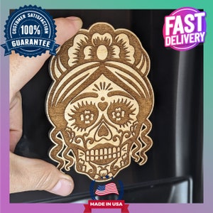 May include: A wooden cutout of a sugar skull with floral details and a headband. The skull is brown and the flowers are white. The cutout is on a black background. "Made in USA" is printed below the cutout.
