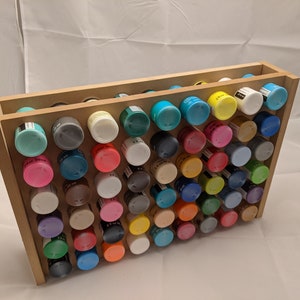 Puede incluir: Un estante de almacenamiento de pintura de madera con dos estantes que sostienen filas de peque&ntilde;os frascos de pintura de varios colores, incluidos azul, verde, rojo, amarillo y blanco.