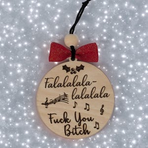 May include: Wooden Christmas ornament with the text "Falalalala-lalala Fuck You Bitch" and musical notes. The ornament has a red bow and a black string for hanging.