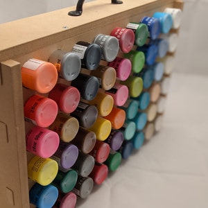 Puede incluir: Una caja de almacenamiento de madera con asa, llena de filas de peque&ntilde;os frascos de pintura. Los frascos est&aacute;n dispuestos en un patr&oacute;n de arco&iacute;ris, con colores como rojo, naranja, amarillo, verde, azul, &iacute;ndigo y violeta.