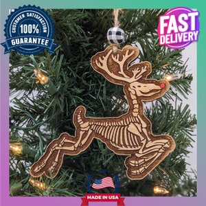 May include: A wooden Christmas ornament shaped like a reindeer skeleton, hanging on a Christmas tree. The reindeer has a red nose and is made in the USA. The ornament is brown with a light brown skeleton design. A black and white checkered bead is at the top.