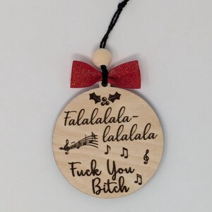 May include: A round wooden Christmas ornament with the text "Falalalala-lalala Fuck You Bitch" and musical notes. It has a red bow and a black string for hanging. The ornament is made of light-colored wood.