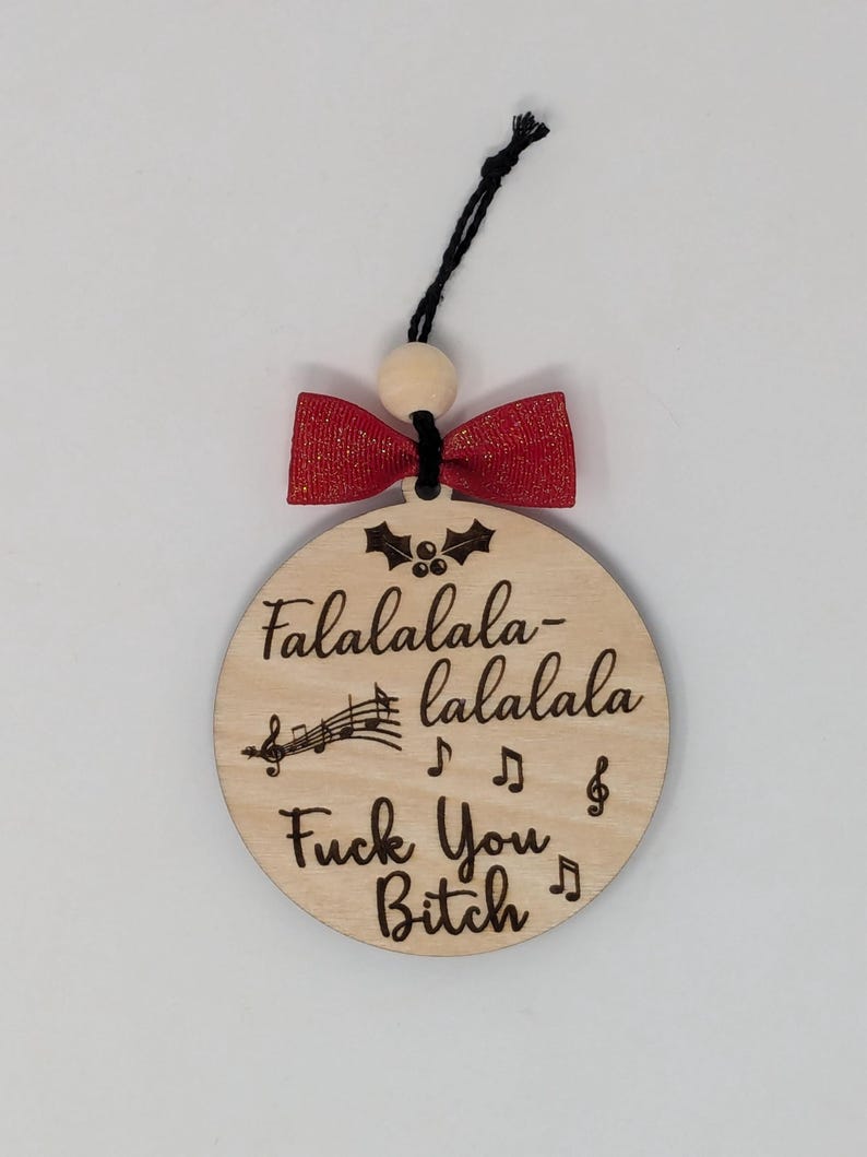 May include: A round wooden Christmas ornament with the text "Falalalala-lalala Fuck You Bitch" and musical notes. It has a red bow and a black string for hanging. The ornament is made of light-colored wood.