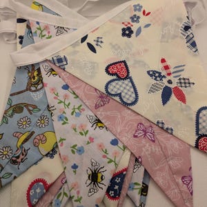 May include: A decorative fabric bunting featuring a variety of patterns, such as bees, butterflies, flowers, and hearts. The pennants are in shades of blue, pink, and cream, connected by a white ribbon. Ideal for party decorations.