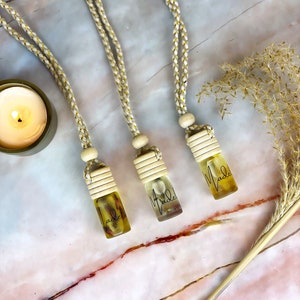 Puede incluir: Tres peque&ntilde;as botellas de vidrio llenas de un l&iacute;quido amarillo y flores secas, suspendidas por cordones trenzados con detalles dorados. Las botellas tienen tapas de madera y la palabra "Made" est&aacute; escrita en ellas. Una vela encendida y plantas secas en el fondo.