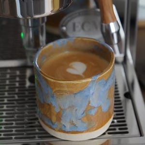 Puede incluir: Una taza de café de cerámica con un latte y arte de espuma en forma de corazón. La taza tiene un esmalte veteado marrón y azul y se encuentra sobre una máquina de espresso de acero inoxidable.