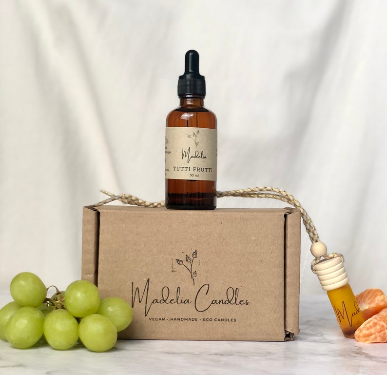 Puede incluir: Una botella de vidrio marr&oacute;n con un cuentagotas negro, etiquetada como "Tutti Frutti", se encuentra sobre una caja marr&oacute;n con el logotipo de "Madelia Candles". Un peque&ntilde;o ambientador para coche cuelga de la caja. Uvas verdes y gajos de naranja en primer plano.