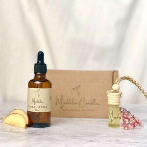 Puede incluir: Una botella de vidrio marr&oacute;n con un cuentagotas, etiquetada como "Floral Apple" y "50 ml", est&aacute; junto a rodajas de manzana. Un peque&ntilde;o ambientador para coche con una parte superior de madera y un cord&oacute;n trenzado cuelga cerca. Una caja marr&oacute;n con el nombre de la marca est&aacute; en el fondo.