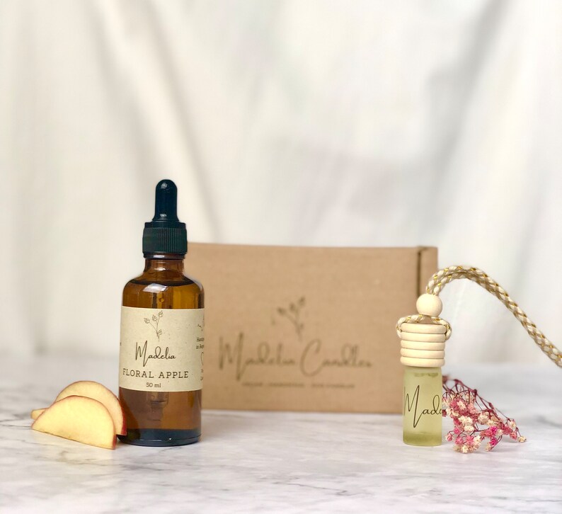 Puede incluir: Una botella de vidrio marr&oacute;n con un cuentagotas, etiquetada como "Floral Apple" y "50 ml", est&aacute; junto a rodajas de manzana. Un peque&ntilde;o ambientador para coche con una parte superior de madera y un cord&oacute;n trenzado cuelga cerca. Una caja marr&oacute;n con el nombre de la marca est&aacute; en el fondo.