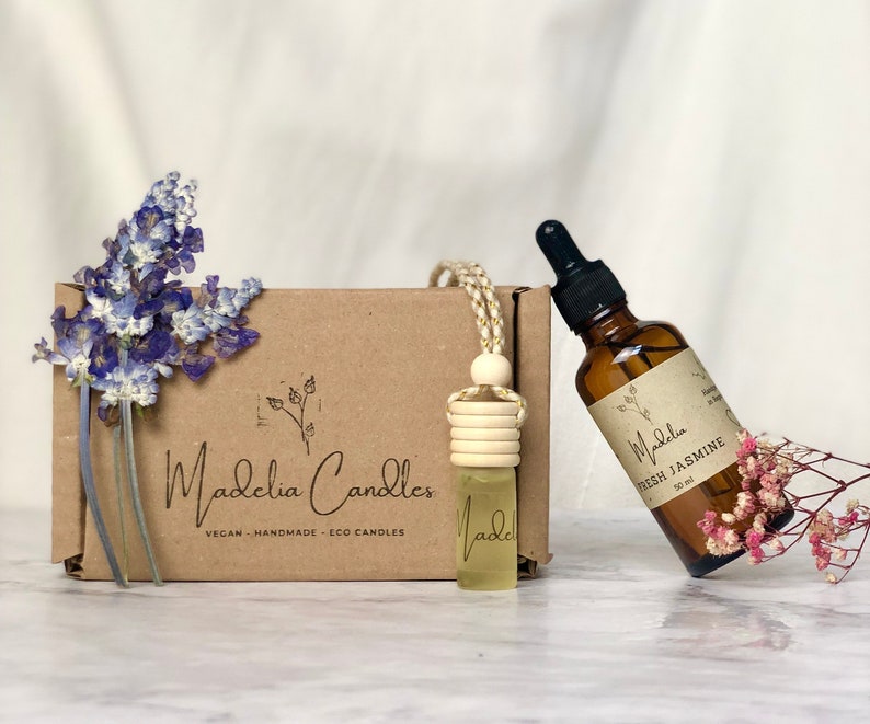 Puede incluir: Una imagen que presenta una caja marr&oacute;n con el texto "Madelia Candles", una peque&ntilde;a botella de vidrio con una cuenta de madera y una botella de vidrio marr&oacute;n etiquetada "Fresh Jasmine". La caja est&aacute; decorada con flores azules.