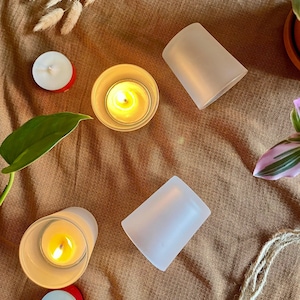 Puede incluir: Vista aérea de velas de té encendidas en portavelas de vidrio transparente, con dos portavelas de vidrio esmerilado y dos velas de té apagadas sobre una superficie de tela marrón. Una hoja verde y una planta están en el marco.