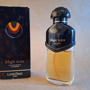Könnte beinhalten: Eine Vintage-Flasche des Parfums "Magie Noire" von Lancôme. Die Parfümflasche ist schwarz mit einem abgerundeten Deckel und einem transparenten Glaskörper. Die Schachtel ist schwarz mit goldenen Akzenten und dem Parfumnamen.