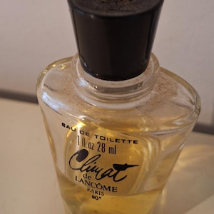 Op de afbeelding: Een kleine, heldere glazen fles "Climat" Eau de Toilette van Lancôme. De fles heeft een zwarte dop en bevat een lichtgele vloeistof. Het etiket luidt "1 fl. oz. 28 ml" en "Paris 80°."