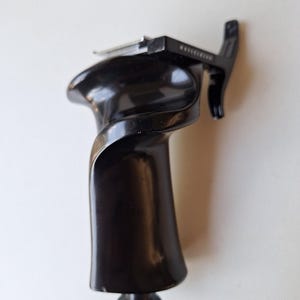 Puede incluir: Un agarre de cámara negro brillante con un diseño curvo. El agarre tiene una perilla texturizada en la base y un componente metálico en la parte superior. La palabra "Hasselblad" está impresa en el componente superior.