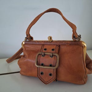 Peut inclure: Un sac à main en cuir marron avec une fermeture à fermoir dorée et une grande boucle en métal. Le sac a une courte poignée et une longue bandoulière.
