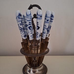 May include: A set of antique knives with white porcelain handles featuring blue floral patterns. The knives are displayed in a silver-toned metal stand with a decorative handle. The stand has a round base and a detailed design.