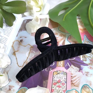 Può includere: Fermaglio per capelli in velluto nero con una grande parte superiore arrotondata e aperta e una fila di denti lungo il fondo. Il fermaglio è su una rivista con un design floreale e il testo "Gucci Flora".