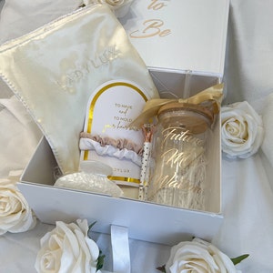 Puede incluir: Una caja de regalo blanca con una inscripción dorada que dice "Bride to Be". La caja está abierta y contiene una máscara de dormir de satén blanco, una diadema para el cabello, un vaso de vidrio transparente con una inscripción dorada que dice "Future Mrs.", un bolígrafo dorado y una tarjeta blanca con el texto "To have and to hold your hair with love".