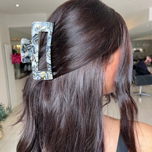 May include: A large, rectangular hair claw with a marbled pattern in shades of gray and white. The claw is made of plastic and has a smooth, glossy finish.