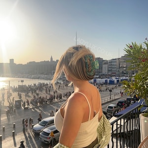 May include: A woman with blonde hair wearing a white tank top and a green and white checkered shirt. She is wearing a large green and blue hair clip. The background shows a cityscape with a harbor and a sunset.