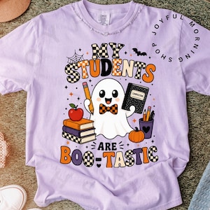 May include: A light purple t-shirt featuring a graphic of a ghost wearing a checkered bowtie, holding a pencil and a composition notebook. The design includes a stack of books, an apple, a pumpkin, and the text My Students Are Boo-Tastic.