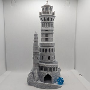 Könnte beinhalten: Ein grauer, 3D-gedruckter Turm mit einem kleineren Turm, der an der Seite befestigt ist. Der Turm hat ein abgerundetes Dach und mehrere Fenster. Der Turm steht auf einem felsigen Sockel mit einem kleinen Höhleneingang. Ein blauer 20-seitiger Würfel befindet sich vor dem Turm.