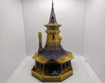 May include: A 3D printed miniature tower with a pointed roof, a chimney, and a small door. The tower is painted in shades of gold and purple, and a blue dice is sitting in front of the door.