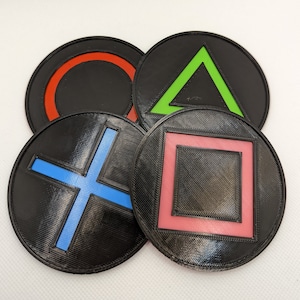 Puede incluir: Cuatro posavasos redondos negros con diferentes símbolos de colores. Los símbolos son un círculo rojo, un triángulo verde, una cruz azul y un cuadrado rosa.