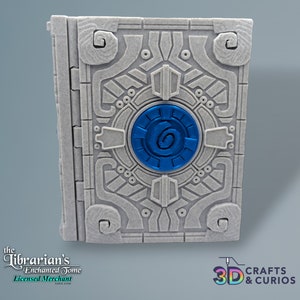 May include: A 3D printed, grey, intricately detailed book with a blue circular design on the front cover. The book is titled "The Librarian's Enchanted Tome" and is a licensed merchant product.