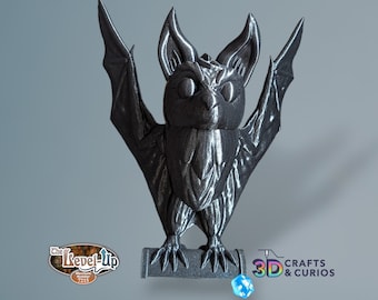 May include: A 3D printed black bat figurine with wings outstretched, standing on a small book. The bat has a detailed, textured surface and a cute, cartoonish face. The book has the text "3D CRAFTS & CURIOS" printed on it.