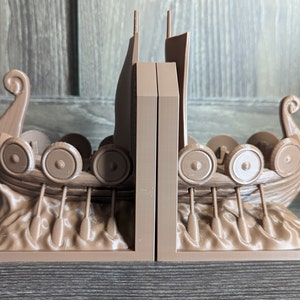 May include: A pair of brown wooden bookends shaped like Viking ships with shields and oars. The bookends are on a wooden surface.