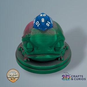 Puede incluir: Una figurita de rana impresa en 3D de color verde y rojo con un dado azul de 20 caras en la parte superior. El dado muestra un 19.