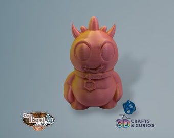 May include: A pink 3D printed monster figurine with a toothy grin, wearing a collar with a hexagonal emblem. The monster has large eyes and a spiky head. A blue 20-sided die is in the foreground.
